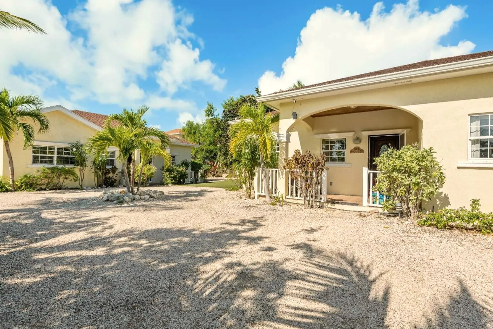 Sunlit gravel courtyard framed by palms, with both cream-coloured villas under a bright blue sky