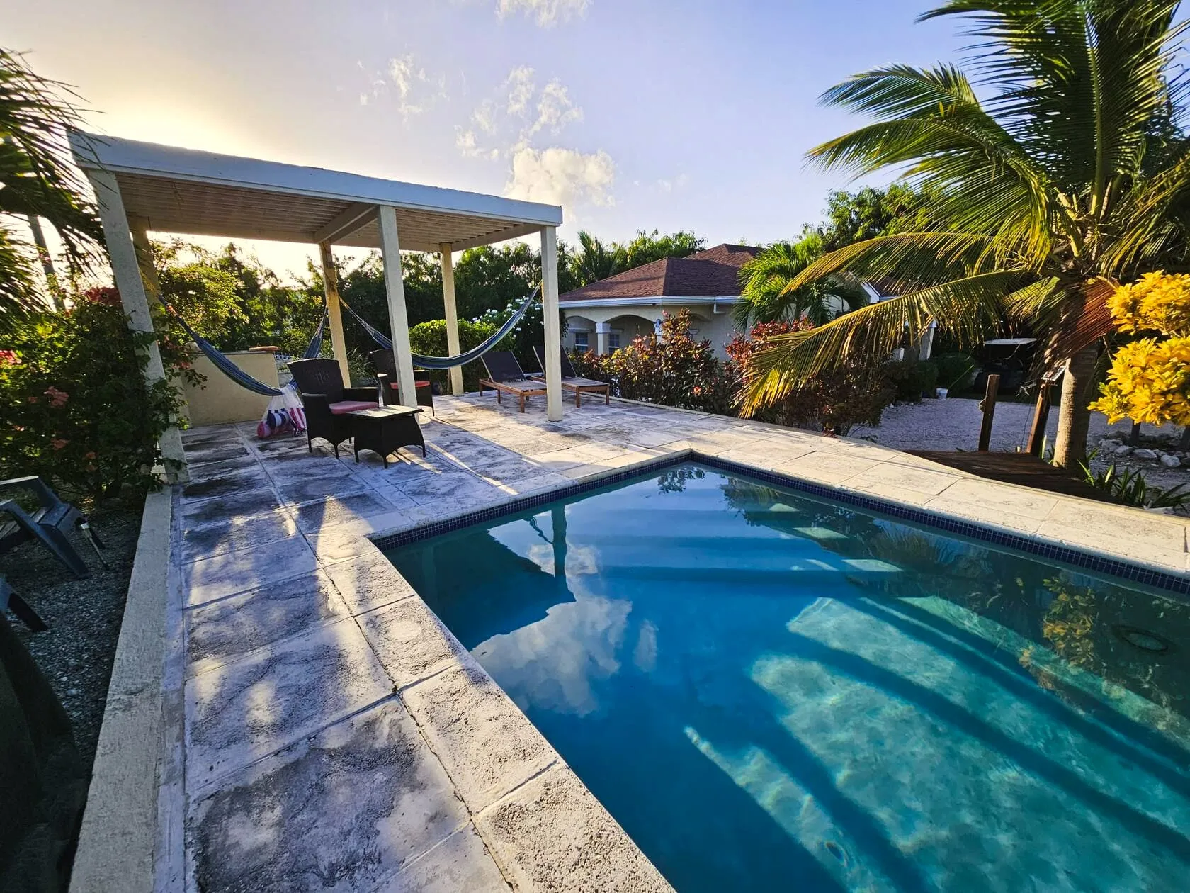 Golden-hour view across the glassy pool toward the pergola, hammocks, loungers, and the villa beyond