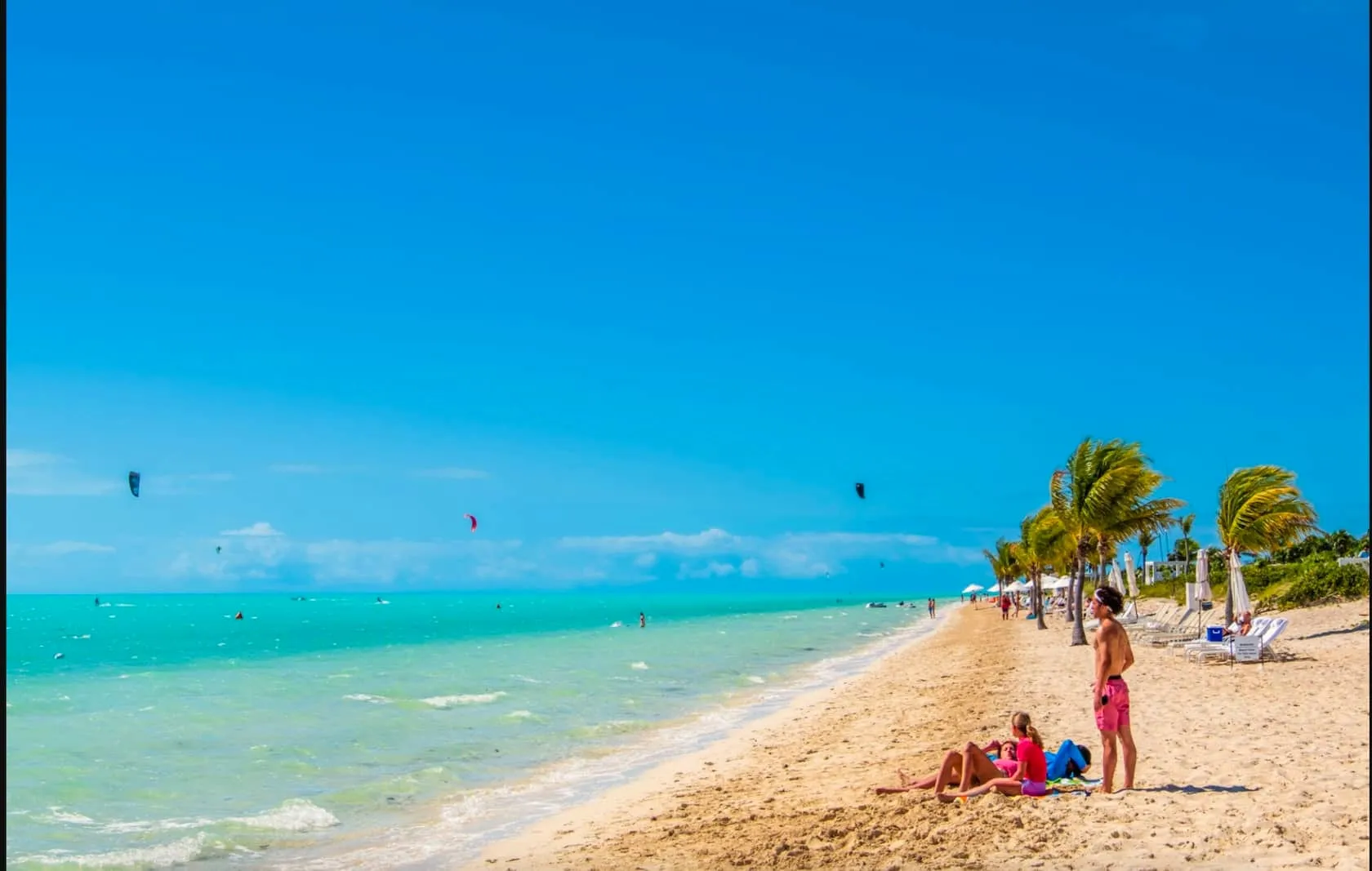 Long Bay Beach with kiteboarders over the turquoise flats and sunbathers on the white sand